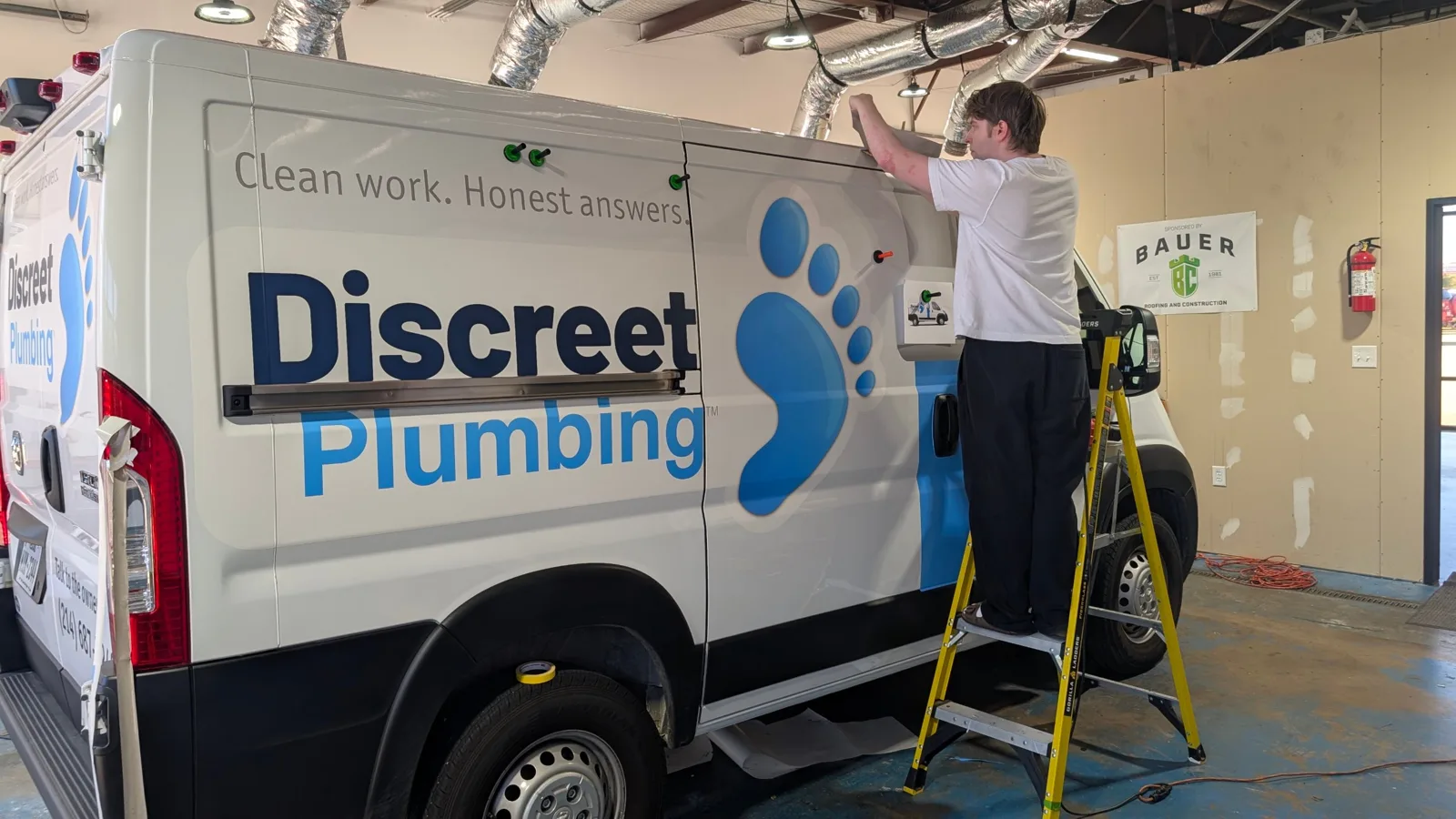 WrapMasters installer applying the Discreet Plumbing partial wrap to a Ram ProMaster in the climate-controlled Denton install bay
